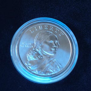 May include: A silver-colored coin encased in a clear plastic protective capsule. The coin features the word LIBERTY at the top and the phrase IN GOD WE TRUST on the left. It depicts a person carrying a small child on their back.
