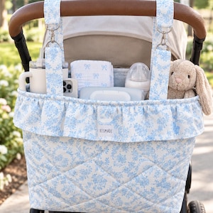 May include: A quilted stroller organizer bag featuring a blue and white floral pattern with a ruffled top edge. The bag hangs from the stroller handle with two straps and metal clips. A small white label reads KESMADE.