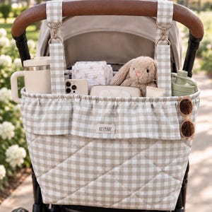 May include: A quilted beige and white gingham stroller organizer bag featuring a ruffled top edge, two hanging straps with gold-tone metal clips, and a small rectangular fabric label with the text KESMADE centered on the front exterior.