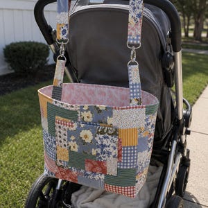 May include: A rectangular fabric tote bag featuring a patchwork design of floral, gingham, and striped patterns in shades of blue, yellow, green, and red. The bag includes two matching fabric straps with silver metal clips for attachment.