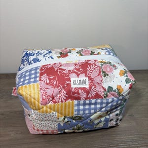 May include: A quilted zippered pouch featuring a patchwork design of various floral and gingham patterns in red, blue, yellow, and white. A white rectangular fabric label with the black text KESMADE is sewn onto the front center of the pouch.
