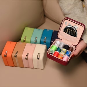 May include: A collection of colorful sewing kits in various shades, including orange, tan, pink, and cream. One kit is open, revealing spools of thread, needles, and small scissors. The kits are arranged on a neutral surface.