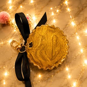 May include: A circular gold-colored charm with a scalloped edge, featuring a metallic swirled finish. It is attached to a black grosgrain ribbon bow and a gold-tone metal lobster clasp. A small pink pom-pom and a clear bead are also attached.