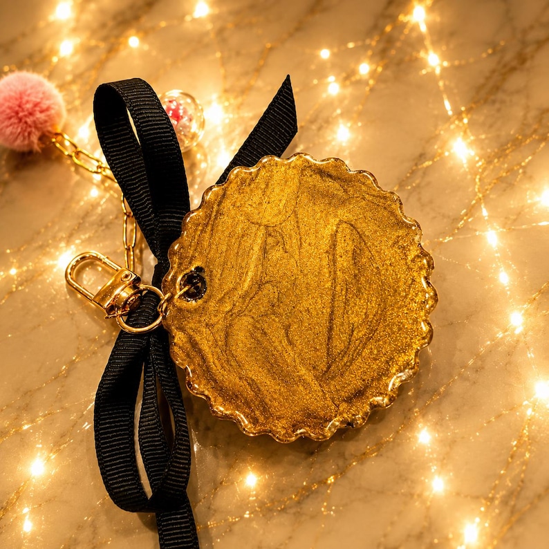 May include: A circular gold-colored charm with a scalloped edge, featuring a metallic swirled finish. It is attached to a black grosgrain ribbon bow and a gold-tone metal lobster clasp. A small pink pom-pom and a clear bead are also attached.