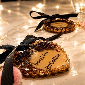 May include: Two heart-shaped resin ornaments with gold glitter edges and a smooth tan center. Each ornament features the black text "Borei Pri HaCoffee," a black ribbon bow at the top, and a small pink pom-pom attached to a gold metal jump ring.
