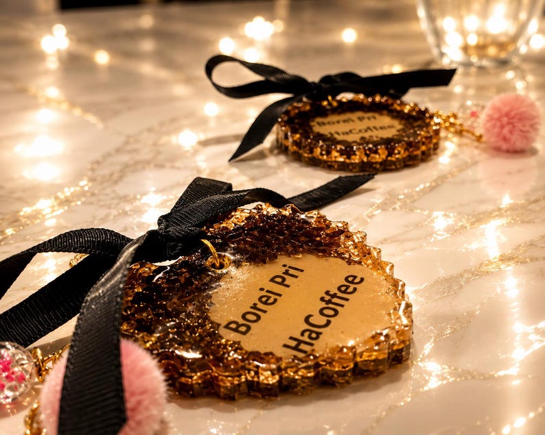 May include: Two heart-shaped resin ornaments with gold glitter edges and a smooth tan center. Each ornament features the black text "Borei Pri HaCoffee," a black ribbon bow at the top, and a small pink pom-pom attached to a gold metal jump ring.