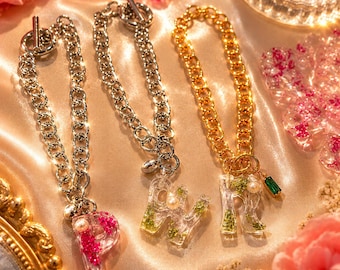 May include: Three chain bracelets with toggle clasps. Two silver-toned chains feature clear resin letter charms P and W, filled with pink and green botanical elements and small pearls. One gold-toned chain features a clear resin letter charm R with green elements, a pearl, and a small green rectangular crystal.