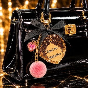 May include: A glossy black handbag featuring a gold-toned clasp and a decorative charm set. The charm includes a black ribbon bow, a pink pom-pom, a small heart-shaped pendant, and a gold glitter leaf tag with the text Borei Pri HaCoffee.