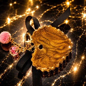 May include: A heart-shaped keychain featuring a shimmering gold resin finish with a scalloped edge. It is attached to a black grosgrain ribbon bow, a gold-toned metal lobster clasp, a small pink pom-pom, and a clear glass bead containing pink flowers.