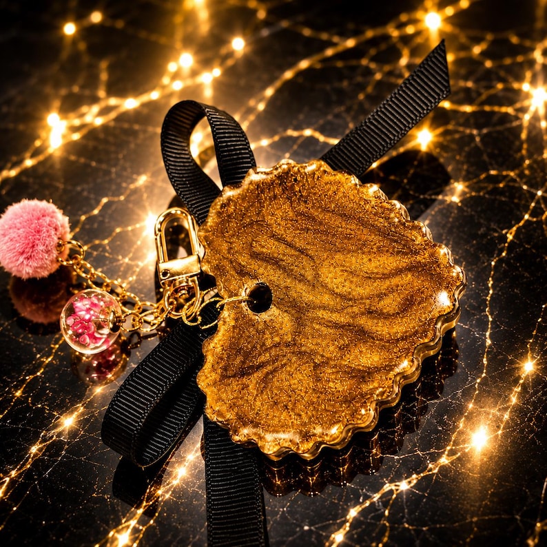 May include: A heart-shaped keychain featuring a shimmering gold resin finish with a scalloped edge. It is attached to a black grosgrain ribbon bow, a gold-toned metal lobster clasp, a small pink pom-pom, and a clear glass bead containing pink flowers.