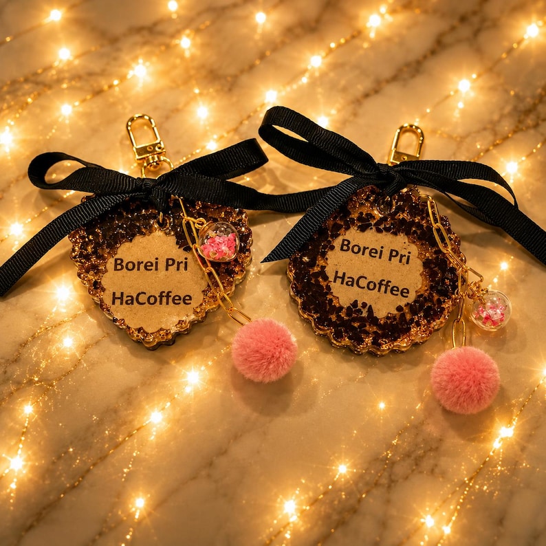 May include: Two keychains featuring a heart-shaped and a circular resin charm with dark brown flecks and a central beige insert printed with the text Borei Pri HaCoffee. Each includes a black ribbon bow, a gold clasp, and a pink pom-pom charm.