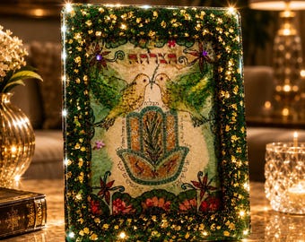 May include: A rectangular picture frame with a dark green and gold glitter border containing an illustration of two birds facing each other above a hamsa hand symbol. The artwork features Hebrew text, floral motifs, and small purple accents.