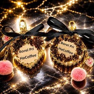 May include: Two circular keychains featuring gold-tone metal hardware, black ribbon bows, and pink faux fur pom-poms. Each keychain has a central beige scalloped tag printed with the text Borei Pri HaCoffee, surrounded by sparkling amber-colored crystals.