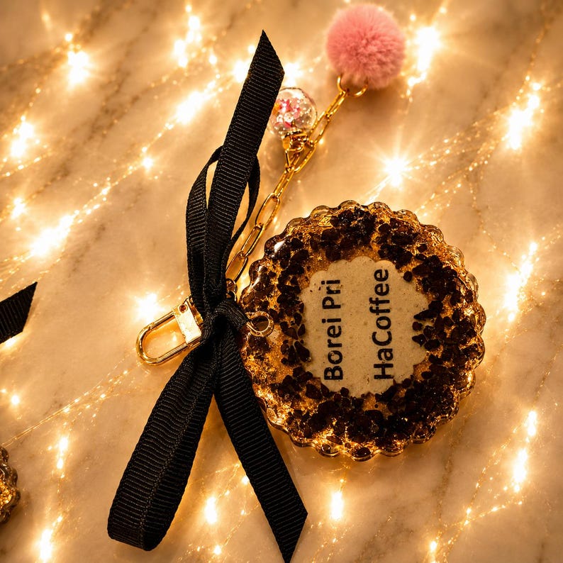 May include: A circular keychain featuring a textured, golden-brown resin base with dark brown flecks and the text "Borei Pri HaCoffee" printed in black. It includes a gold-toned metal chain, a black ribbon bow, and a small pink pom-pom charm.