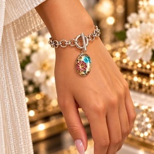 May include: A silver-toned chain link bracelet featuring a toggle clasp and a single oval-shaped pendant. The pendant contains a clear resin inlay with embedded blue and pink floral accents, along with small gold-colored flecks throughout.