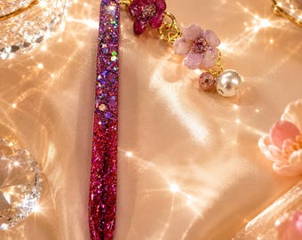 May include: A pen with a glittery ombre finish transitioning from deep purple to vibrant magenta. Attached to the top is a gold-toned chain featuring a dark pink flower, a light pink flower, a small faceted bead, and a single white pearl bead.
