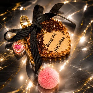 May include: A heart-shaped keychain featuring a brown, textured surface with the text Borei Pri HaCoffee. It is adorned with a black ribbon bow, a gold metal clasp, a small heart charm filled with pink glitter, and a soft, round pink pom-pom.