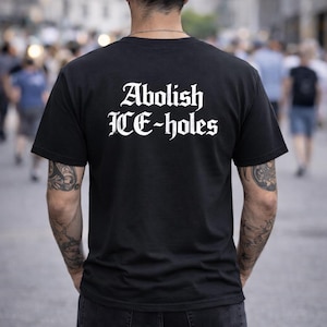 Peut inclure: T-shirt noir avec l'inscription "Abolish ICE-holes" en blanc, en police gothique. Le t-shirt à manches courtes est porté par une personne tatouée aux bras. L'arrière-plan est flou, suggérant un environnement extérieur.