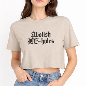 Peut inclure: Un crop top beige avec l'inscription "Abolish ICE-holes" en lettres gothiques noires. Le t-shirt a des manches courtes et un col rond. Le modèle porte un jean bleu.