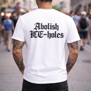 Peut inclure: T-shirt blanc avec l'inscription "Abolish ICE-holes" imprimée en lettres gothiques noires au dos. Le t-shirt est porté par une personne tatouée sur les bras, debout dans une foule.