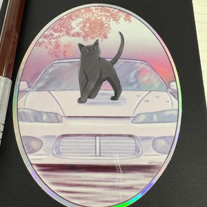 Op de afbeelding: Ovale sticker met een grijze kat op de motorkap van een witte auto. De achtergrond toont een roze en paarse lucht met een boom en een vlinder. De sticker heeft een holografische rand.