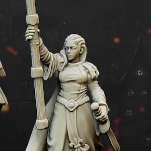 May include: A white painted miniature figurine of a woman wearing a long robe and holding a staff topped with a fleur-de-lis. The figurine is standing on a round base.