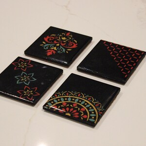 Puede incluir: Cuatro posavasos cuadrados negros con diseños florales y geométricos coloridos. Cada posavasos presenta un patrón único en rojo, amarillo y turquesa. Los posavasos están dispuestos sobre una superficie blanca.