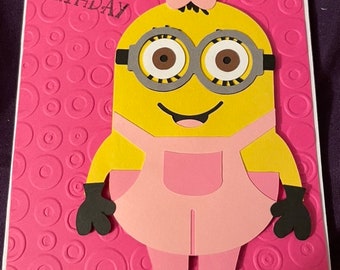 May include: A pink and yellow birthday card featuring a female Minion character with a pink bow in her hair. The card has a pink background with a circle pattern and the text "Happy Birthday".