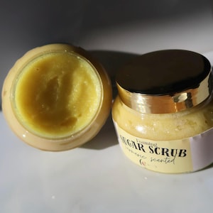 Puede incluir: Dos tarros de exfoliante de azúcar espumoso con aroma a cúrcuma. Un tarro está abierto, revelando el exfoliante amarillo. El otro tarro está cerrado con una tapa dorada. La etiqueta dice "Foaming Sugar Scrub turmeric scented".