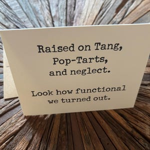 May include: A white greeting card with black typewriter-style text that reads: Raised on Tang, Pop-Tarts, and neglect. Look how functional we turned out. The card is folded and stands upright, displaying the printed message clearly on the front.