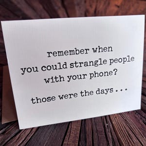May include: A folded greeting card with the text "remember when you could strangle people with your phone? those were the days..." printed in black. The card is off-white and set against a dark wood surface.