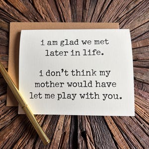 May include: A white greeting card with black typewriter-style text that reads: i am glad we met later in life. i don't think my mother would have let me play with you. A gold-colored pen rests diagonally across the card, which sits on a brown envelope.