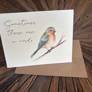 May include: A white greeting card featuring a watercolor illustration of a small bird perched on a brown branch. The text "Sometimes there are no words" is printed in black cursive script on the left side. A brown envelope is placed underneath.