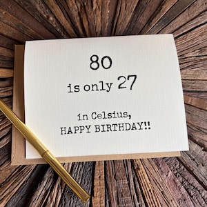 May include: A cream-colored greeting card with black text that reads 80 is only 27 in Celsius, HAPPY BIRTHDAY!!, resting on a brown envelope with a gold-toned pen placed diagonally across the front of the card.