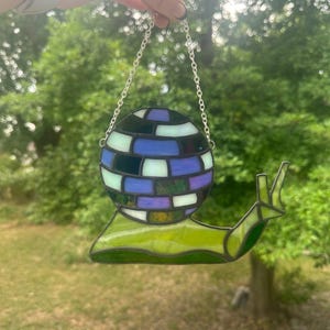 Può includere: Un ornamento a forma di lumaca in vetro colorato con un guscio a palla da discoteca. Il corpo della lumaca è verde e il guscio è un mosaico di pezzi di vetro blu, viola, neri e bianchi. È appeso a una catena d'argento, ideale per una decorazione da finestra.