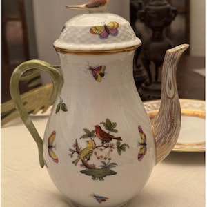 May include: A white porcelain teapot featuring a lid topped with a small bird figurine. The body is decorated with colorful butterflies and a central illustration of two birds perched on a branch. It has a green handle and a textured spout.