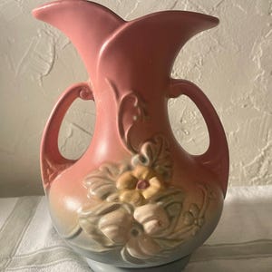 May include: A vintage pink vase with a flared top and two handles. The vase features a floral design in shades of yellow, cream, and green. The base of the vase is a light blue color. The vase is likely made of ceramic.