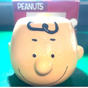 May include: A ceramic mug shaped like Charlie Brown's head, with a tan face, black hair, and a smiling expression. The mug has a rounded shape and is set against a background with the word "PEANUTS".