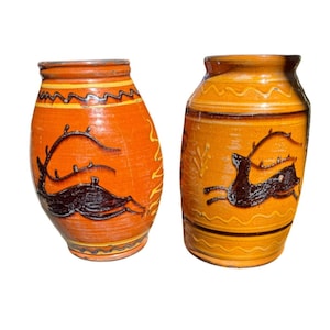 May include: Two ceramic vases with a glossy orange and yellow glaze. Each vase features a dark brown relief design of a leaping deer with antlers. The surfaces are decorated with horizontal wavy lines and bands, showcasing a rounded, bulbous shape.