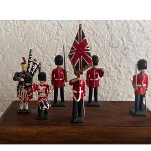 May include: A set of six miniature metal figurines depicting British soldiers in red tunics and black bearskin hats, including a bagpiper, a drummer, and a flag bearer holding a Union Jack, all mounted on a rectangular wooden display base.