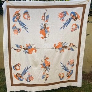May include: A vintage white tablecloth with a brown border and colorful floral designs. The floral patterns include orange, blue, and brown elements. The tablecloth is hanging outside on a clothesline.
