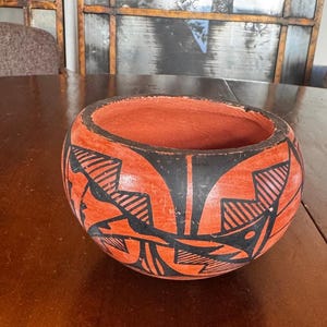 May include: A round, terracotta-colored clay bowl featuring a wide opening and a matte finish. The exterior is decorated with bold, black geometric patterns, including sharp angular lines and hatched details that wrap around the curved surface.