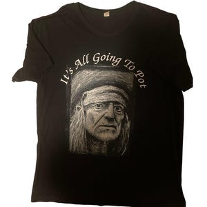 May include: Black t-shirt featuring a detailed grayscale portrait of a man with long hair and a bandana. The text "It's All Going To Pot" is arched above the portrait in white lettering. The shirt has a crew neck.