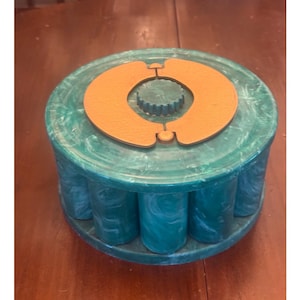 May include: A round, teal and white marbled container with a cylindrical body and a flat lid. The lid features a central, textured, orange, semi-circular dual-piece design with a small, ridged, circular knob in the very center.