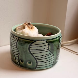 以下が含まれることがあります： 内側と外側が薄緑色のセラミック製ニンニクキーパー。外側は黒い線と3つの小さな穴がある濃い緑色のデザインです。キーパーにはニンニクの頭がいくつか入っています。