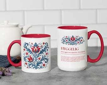 Könnte beinhalten: Zwei weiße Keramikbecher mit roten Innenräumen und Griffen. Ein Becher hat ein florales Design in Rot und Blau. Der andere Becher hat das Wort "HYGGELIG" in Rot mit einem floralen Design darüber. Die Becher stehen auf einer grauen Oberfläche.