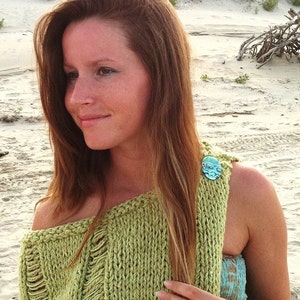 May include: A woman wearing a green knitted sweater with a distressed look. The sweater has a light blue button on the shoulder. She is wearing a light blue tank top underneath the sweater.