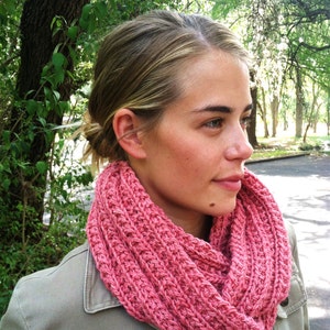 Könnte beinhalten: Eine Frau trägt einen rosafarbenen, gestrickten Infinity-Schal. Der Schal ist aus weichem, dickem Garn gefertigt und hat ein schlichtes, elegantes Design.