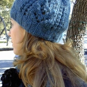 May include: A woman with long, light brown hair is wearing a blue knitted hat with a cable knit pattern. She is also wearing a blue scarf and a black jacket.