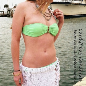 Puede incluir: Una mujer con un top de bikini verde y una falda de crochet blanca. La falda es un patrón de crochet con una abertura lateral. La mujer está de pie en un muelle de madera con un barco en el fondo.